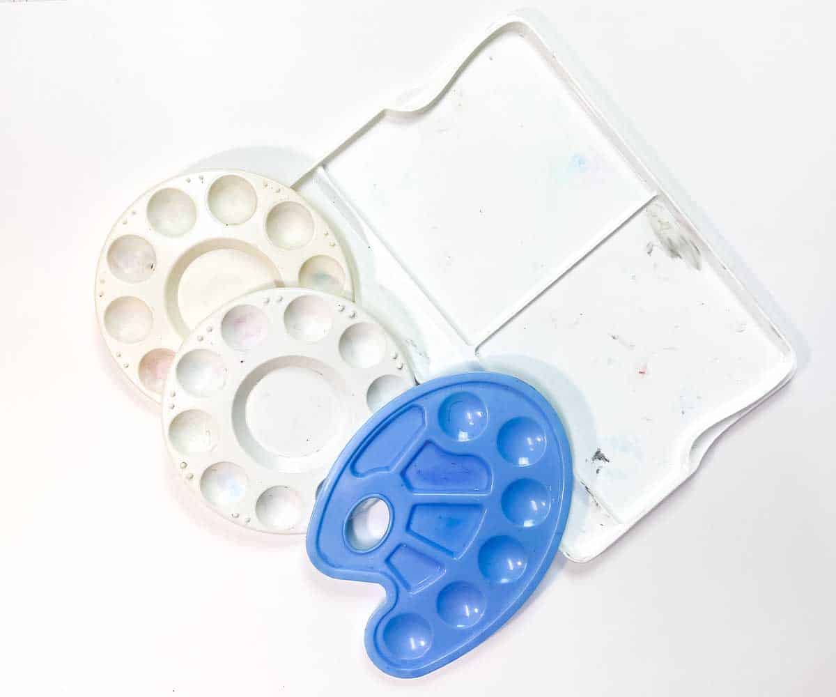 Artist's plastic paint palettes. Two white round palettes with paint wells. One blue palette with paint wells and a thumb hole. One large flat palette divided into two sections.