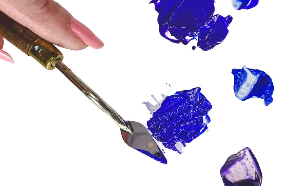 A hand holding a palette knife mixing blue and purple paint.