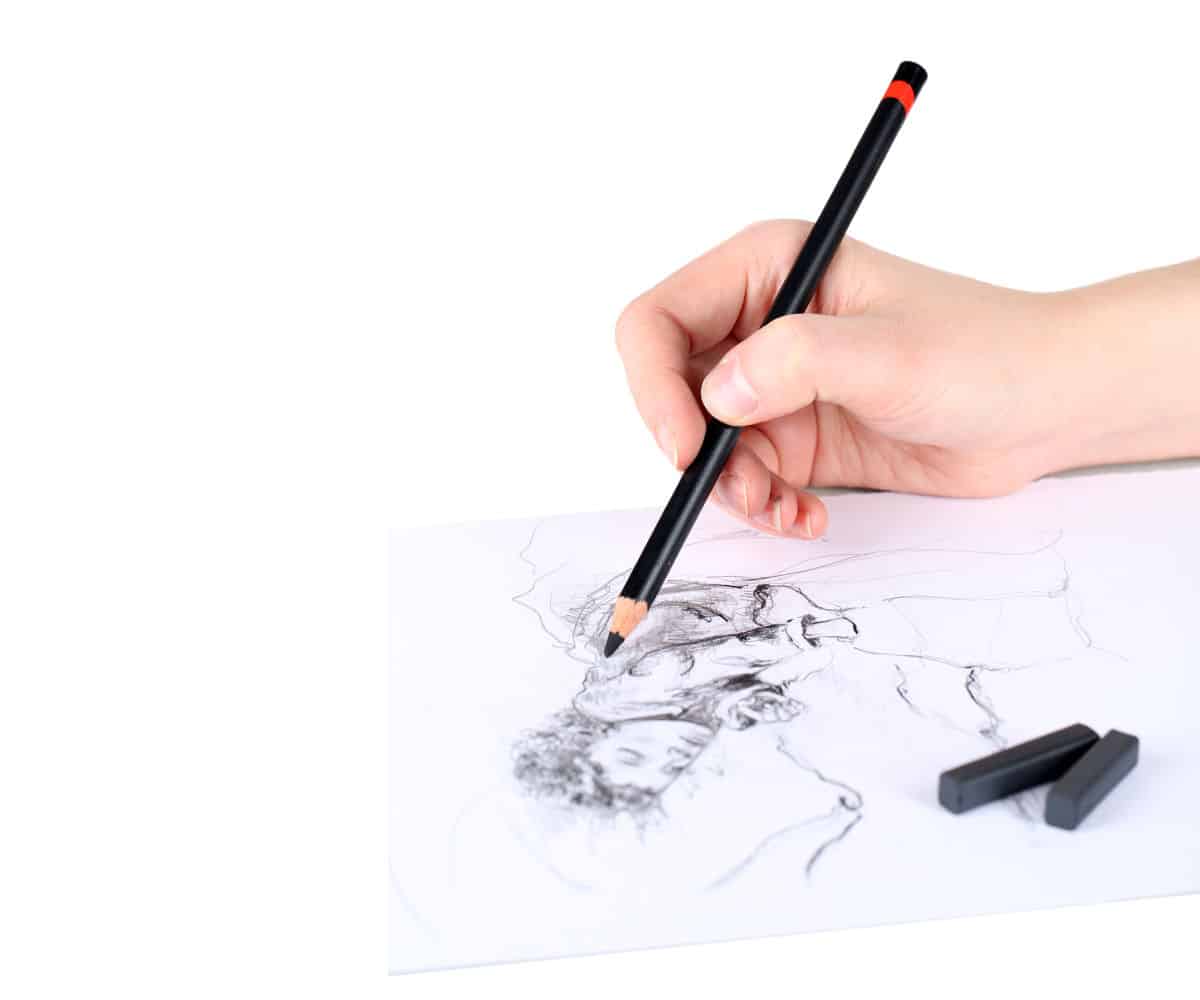 A hand holding a drawing pencil above a drawing in progress with two charcoal sticks on a white background.