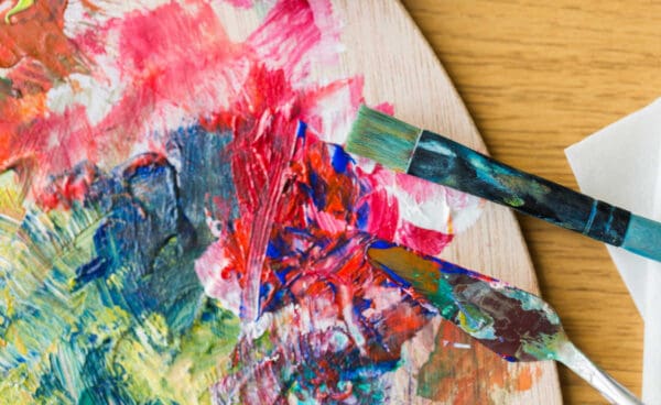 A picture of various colors of thick paint and a palette knife and brush on a wooden painters palette.