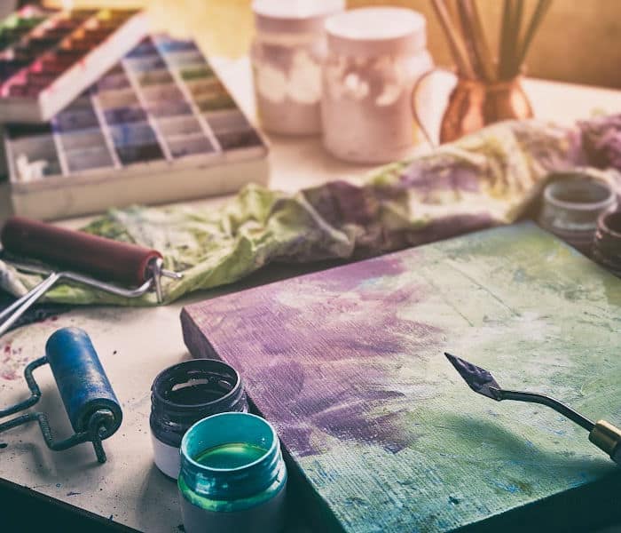 An artists' canvas on a table surrounded by paint pots, brushes, palette knives and other art supplies.