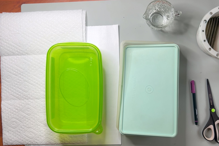 Supplies for making a diy wet palette. Paper towels, palette paper, two plastic containers with lids, a jug of water, a pen, and scissors.